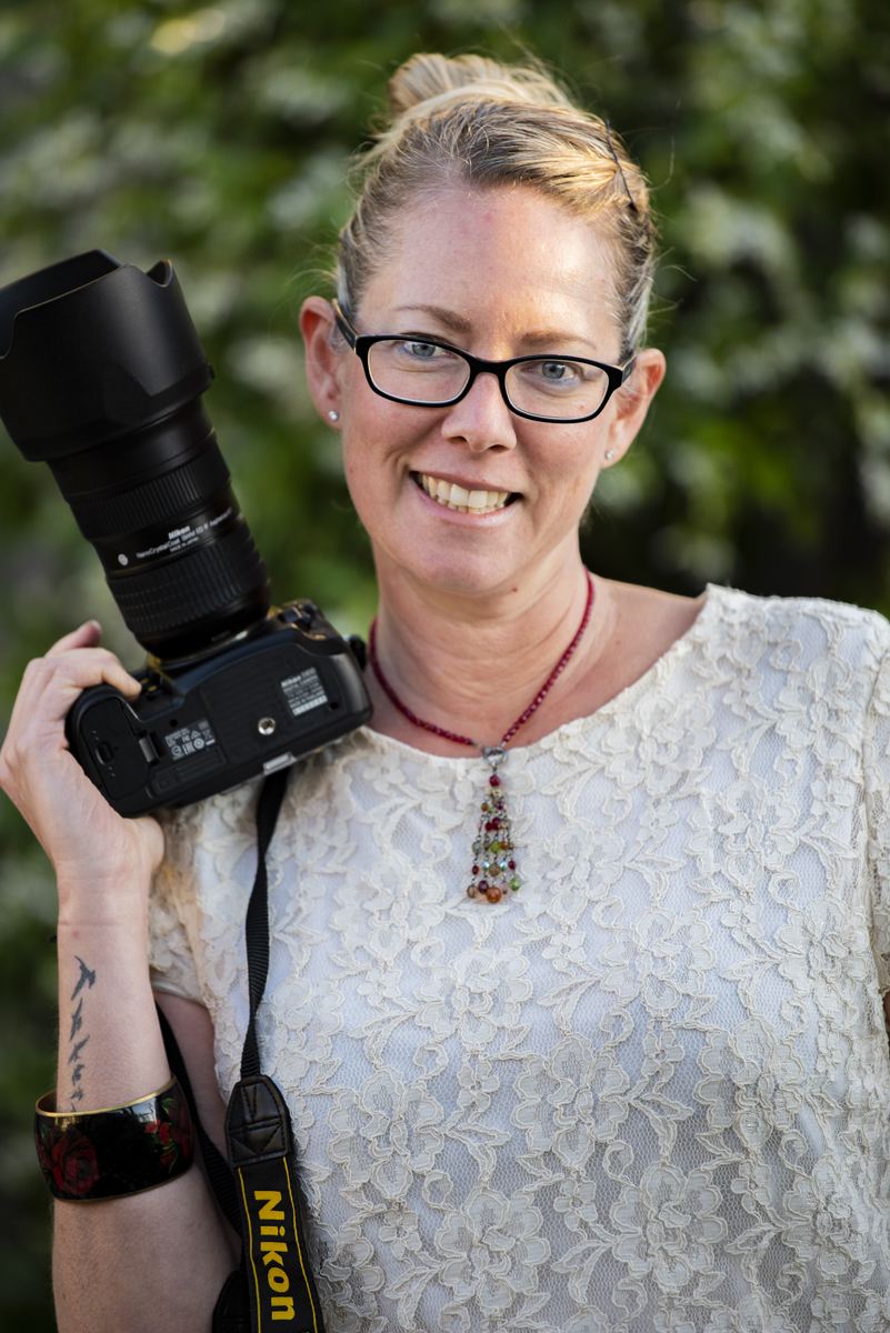 Michelle is standing in front of a leafy green background, holding a camera over her shoulder